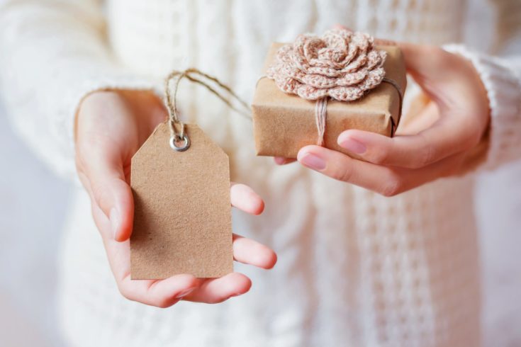 Woman in white knitted sweater holding a present. Gift is packed in craft paper with hand made crocheted flower.Empty tag for your text. Mock up. Example of DIY ways to pack Cristmas and other presents.