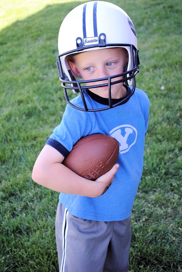 Little boy in blue t-shirt and gray shorts,holding a football and wears a football helmet in an open ground.