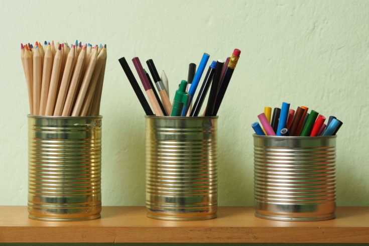 Crayons and pens in waste tin cans standing on a shelf.