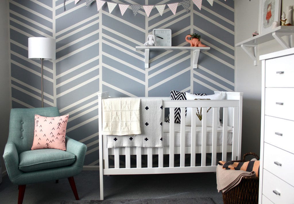 Accent wall with a blue green chair and a pink pillow. A white crib below a wall clock and a white drawer.