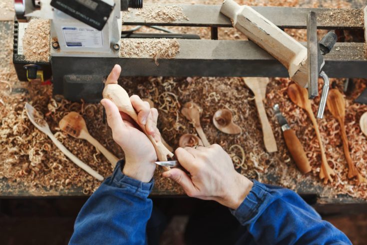 Man's hands in blue jeans working suit carving a wooden spoon with a knife, wooden shavings, spoons and instruments on table at background, close up, woodworking.