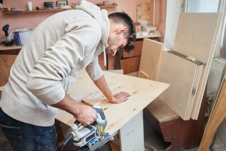 Young man working with electric fretsaw while sawing plywood,