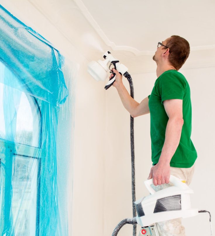 Man painting room walls with paint spray gun while standing on ladder with the best airbrush compressor tank