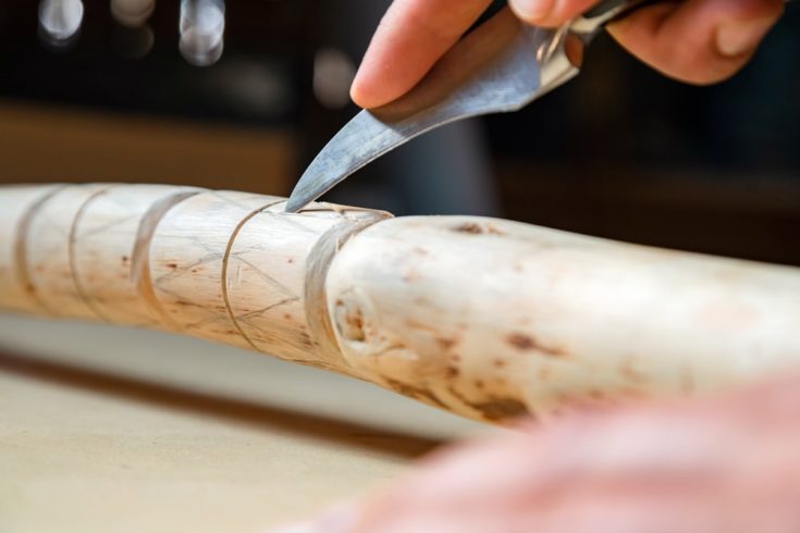 Process of man making wooden walking stick indoors during quarantine. Carving wood stick on the table using knife