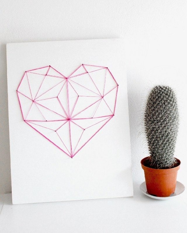 Geometrical heart red string art design on white plain board and potted cactus on his side.