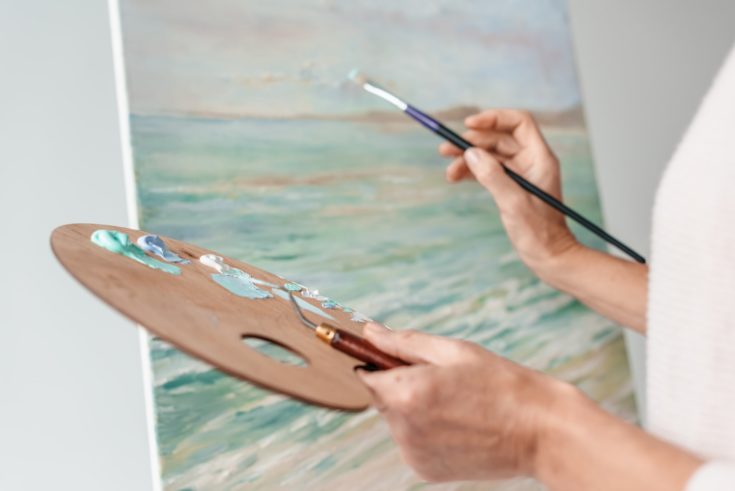 Cropped shot of artist holding palette and paintbrush while painting at easel in art studio