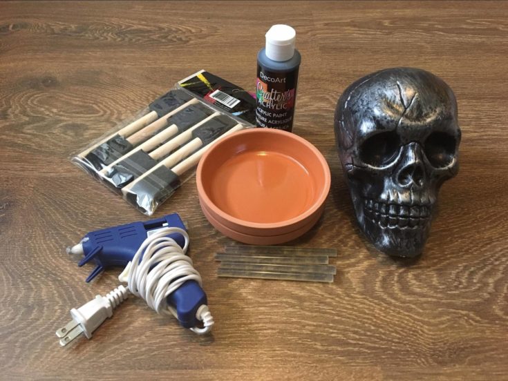 Glue gun + terra cotta + paint + paint brush + plastic skull + glue sticks on the wooden table