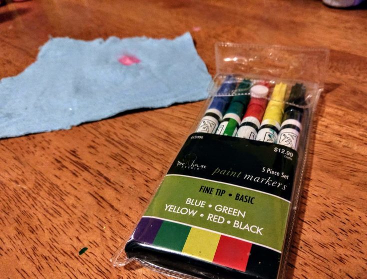 5 colors paint markers on top of a wooden table and blue cloth on the side.