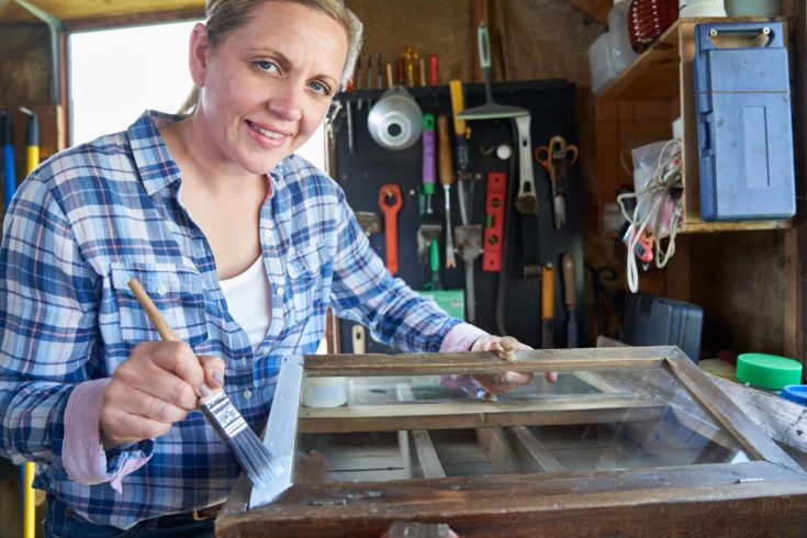 Portrait Of Mature Woman Upcycling Furniture In Workshop At Home Painting Cabinet