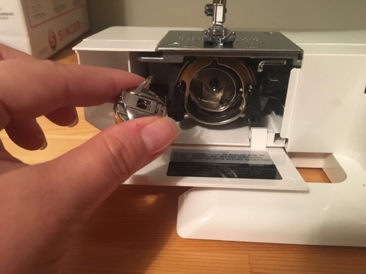 a person's hand holding a bobbin holder removing it from the sewing machine