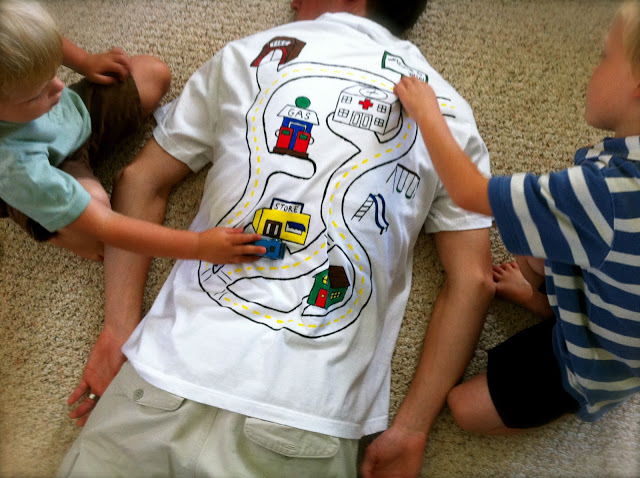 Dad lying on the floor wearing Car Shirt, with 2 boys playing with their car on his back