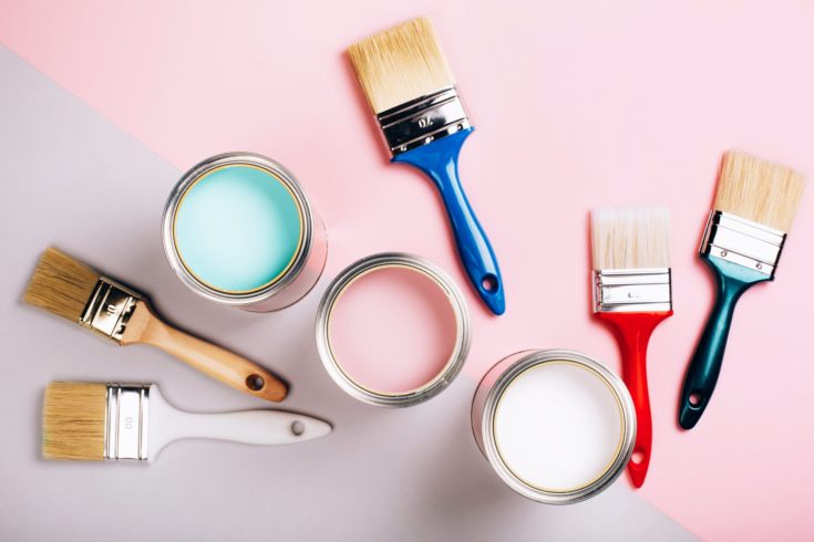 Three open cans of pastel paint with five brushes on pink and grey background. Renovation concept.