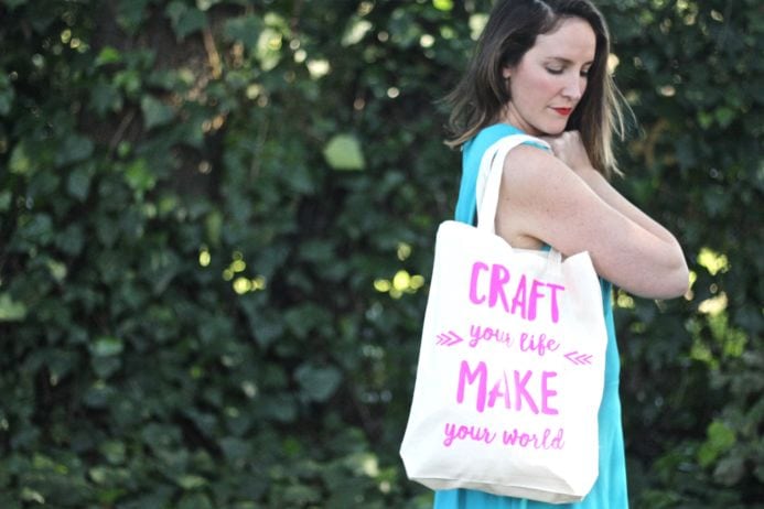 Woman in a light blue dress holding a DIY heat press tote bag with greeny leaves background.