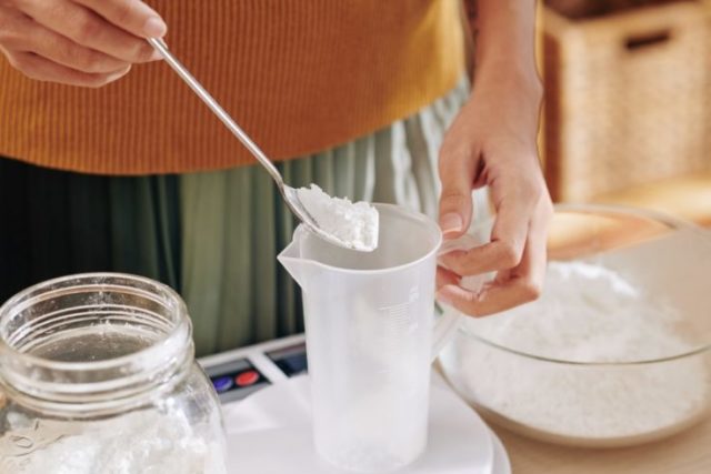 Close-up image of woman putting spoon of lye in plastic jar when making soap at home