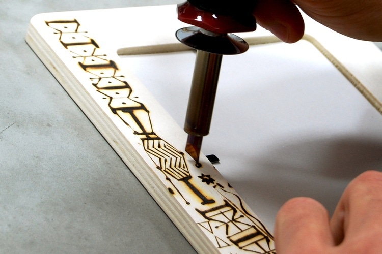 A close up shot of hands holding a wood burning tools to make designs on wood frame.